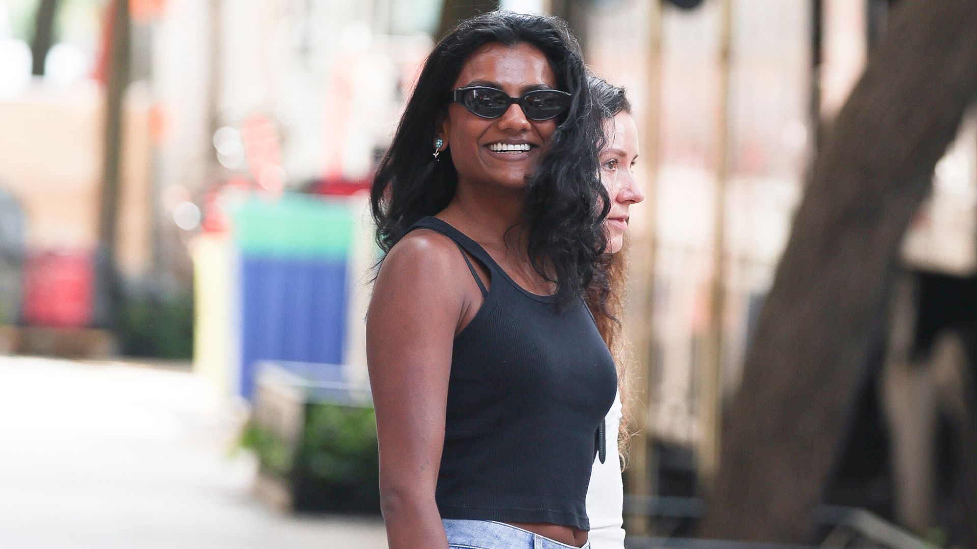 Simone Ashley stands outside wearing a black tank top with blue flared jean and black sunglasses.