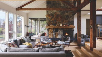 Spacious living room with exposed wooden beams, stone fireplace, burning hearth, built-in log storage, and comfortable gray sectional sofa