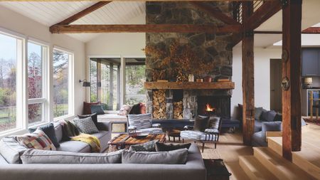 Spacious living room with exposed wooden beams, stone fireplace, burning hearth, built-in log storage, and comfortable gray sectional sofa