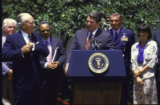 a group of people stand behind ronald reagan with a podium. the men wear suits and sally ride, visible on right, wears a white dress
