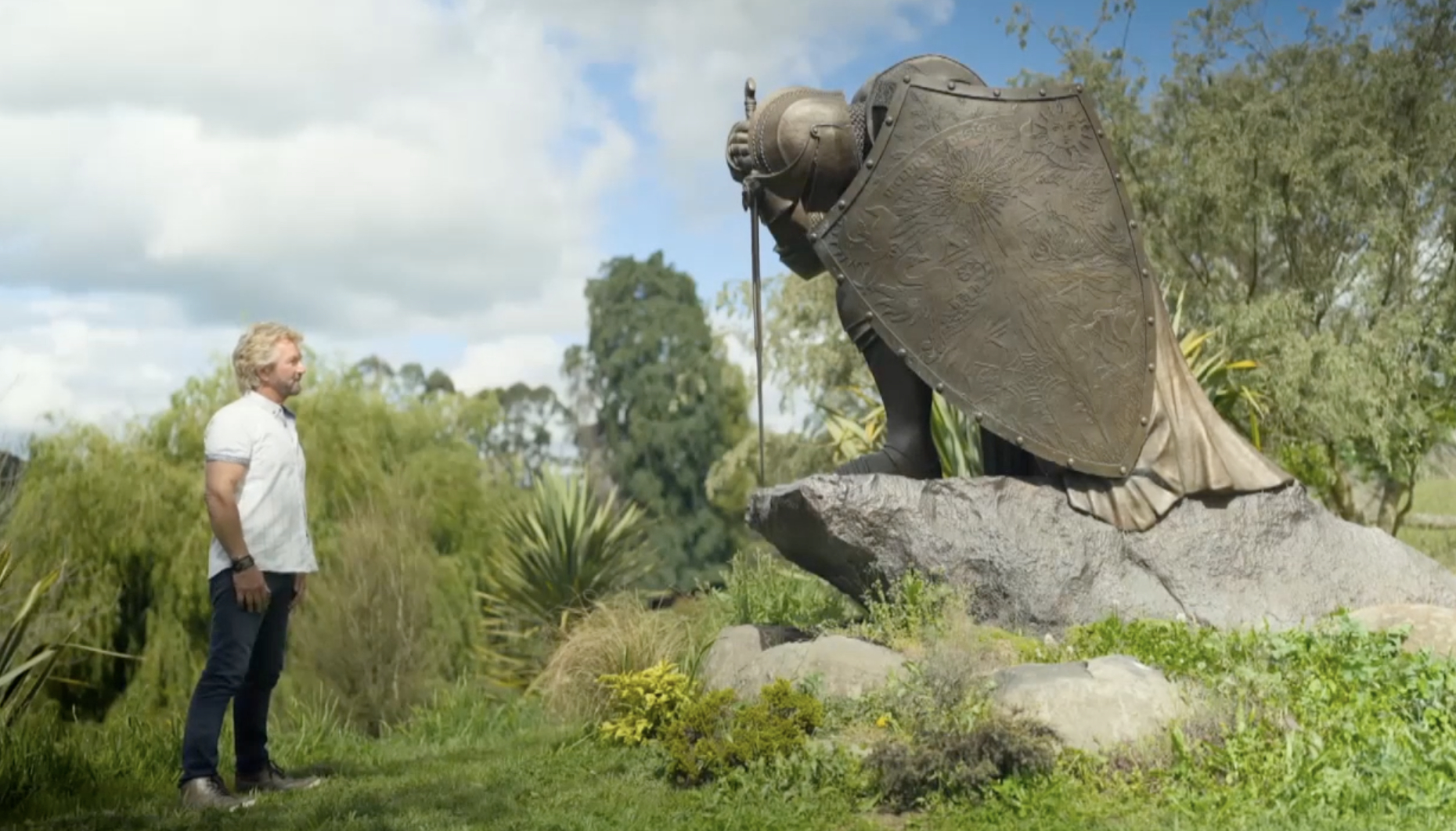 Noel Edmonds in a garden looking at an enormous statue of a warrior kneeling.