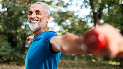 Older male in blue t-shirt holding dumbbells out at shoulder-height in a local park smiling