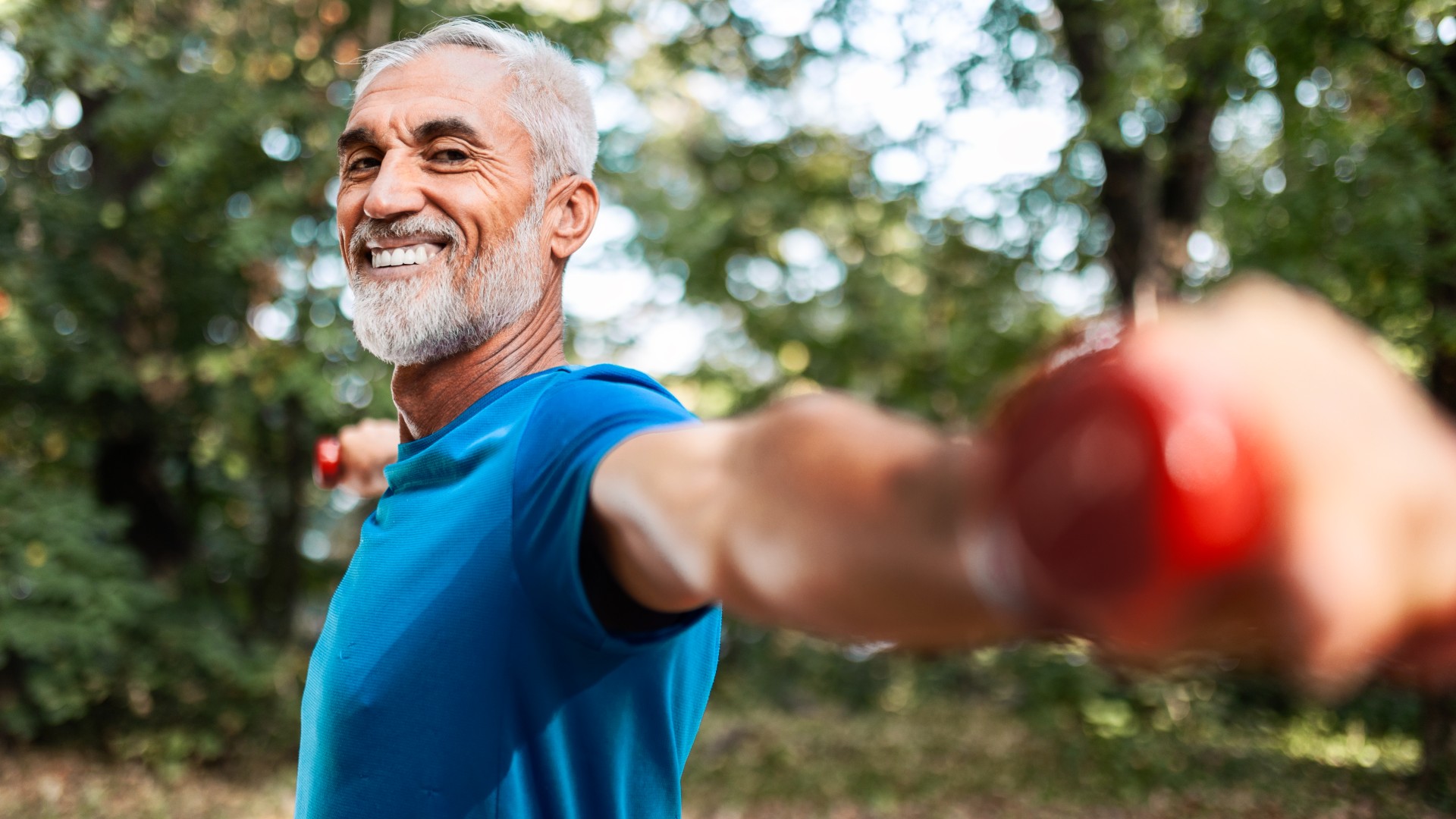 Older male in blue t-shirt holding dumbbells out at shoulder-height in a local park smiling