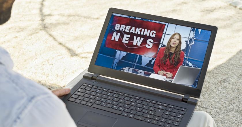 Man watching newscaster presenting breaking news on laptop while sitting outdoors in public park.