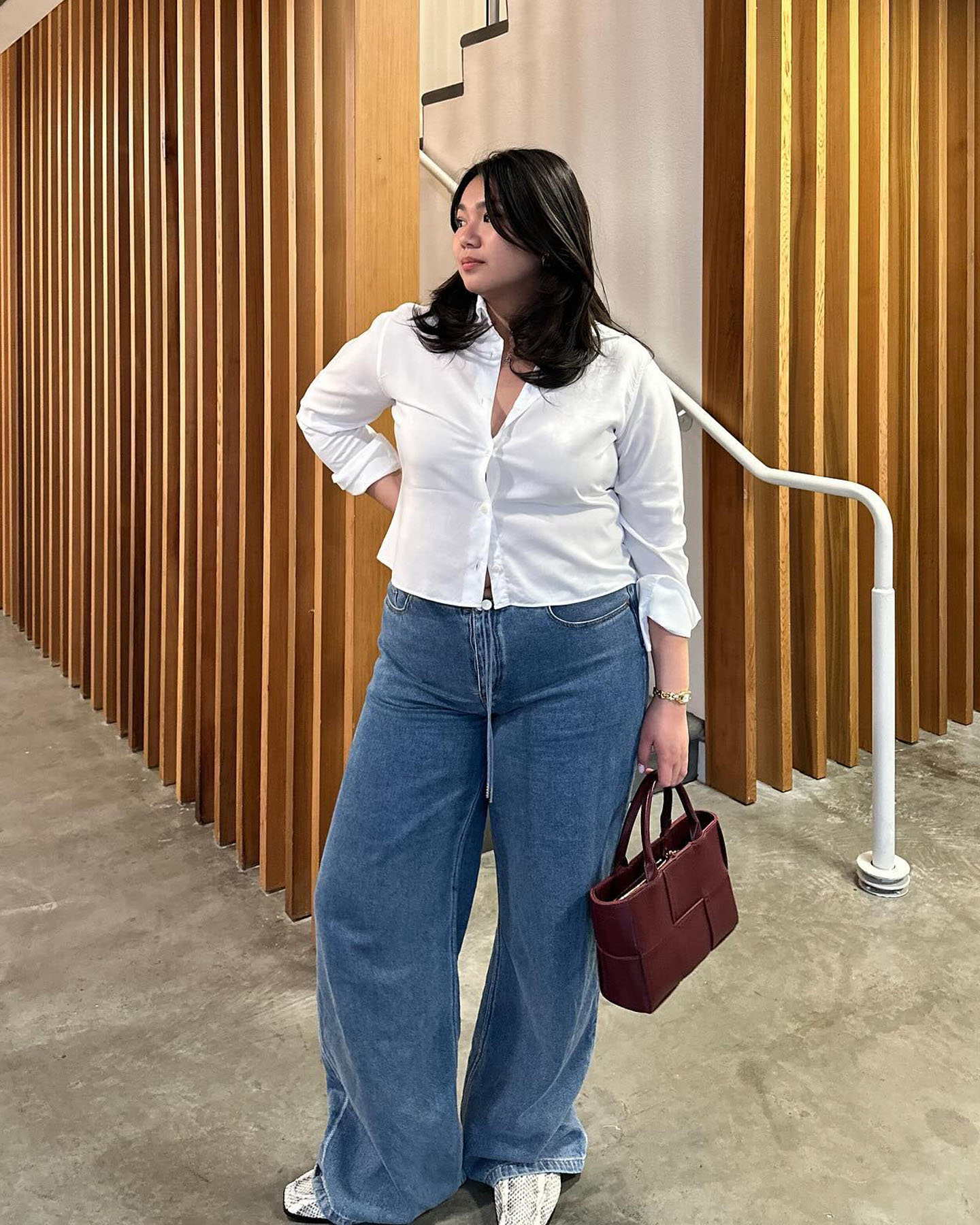 mid-size fashion influencer Marina Torres poses in a white button-down shirt, a burgundy Bottega Veneta bag, wide-leg jeans, and snakeskin shoes