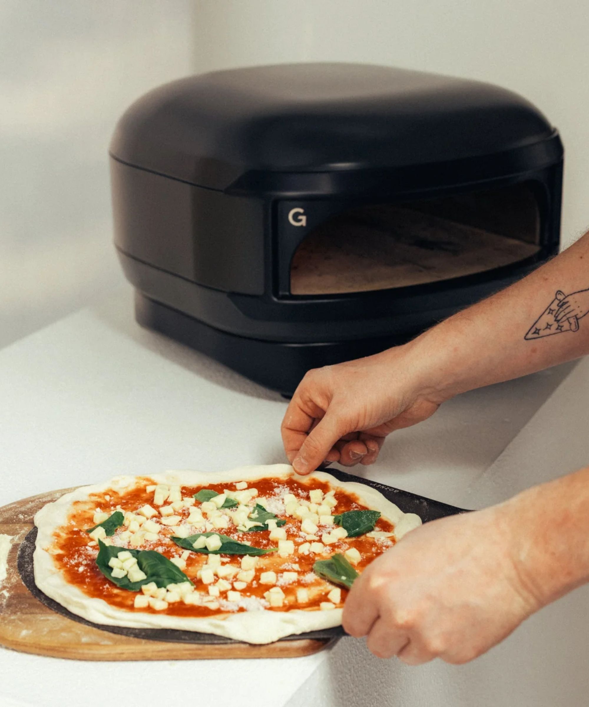 The Gozney Arc Lite slim black pizza oven on a white table, with a mans hands shuffling an uncooked pizza onto a pizza slice in front of it
