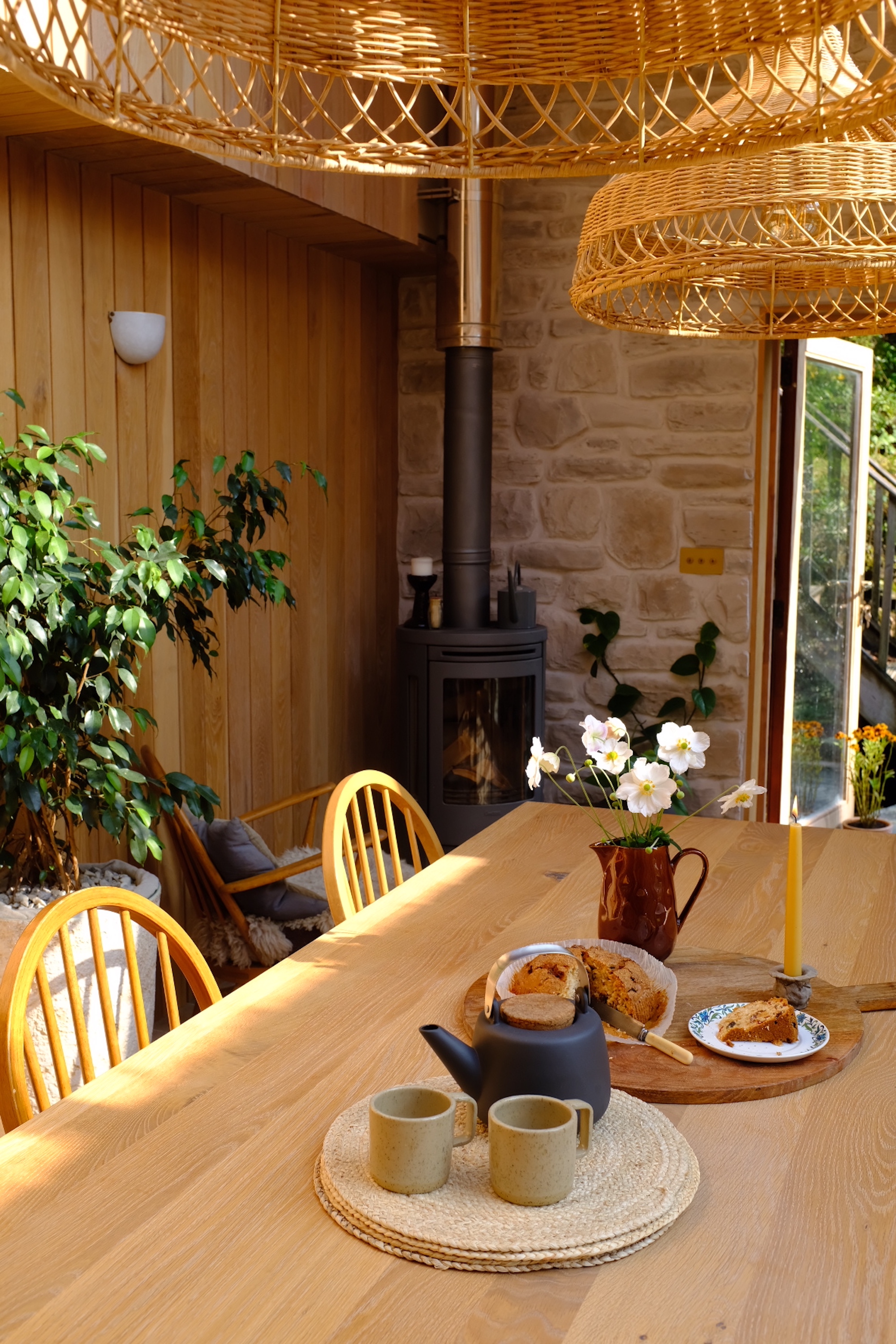 Dining table with a tea set and a jug with flowers on the top, and large rattan pendant lights above - in the background is a small wood burner and a view through to the outdoor space.