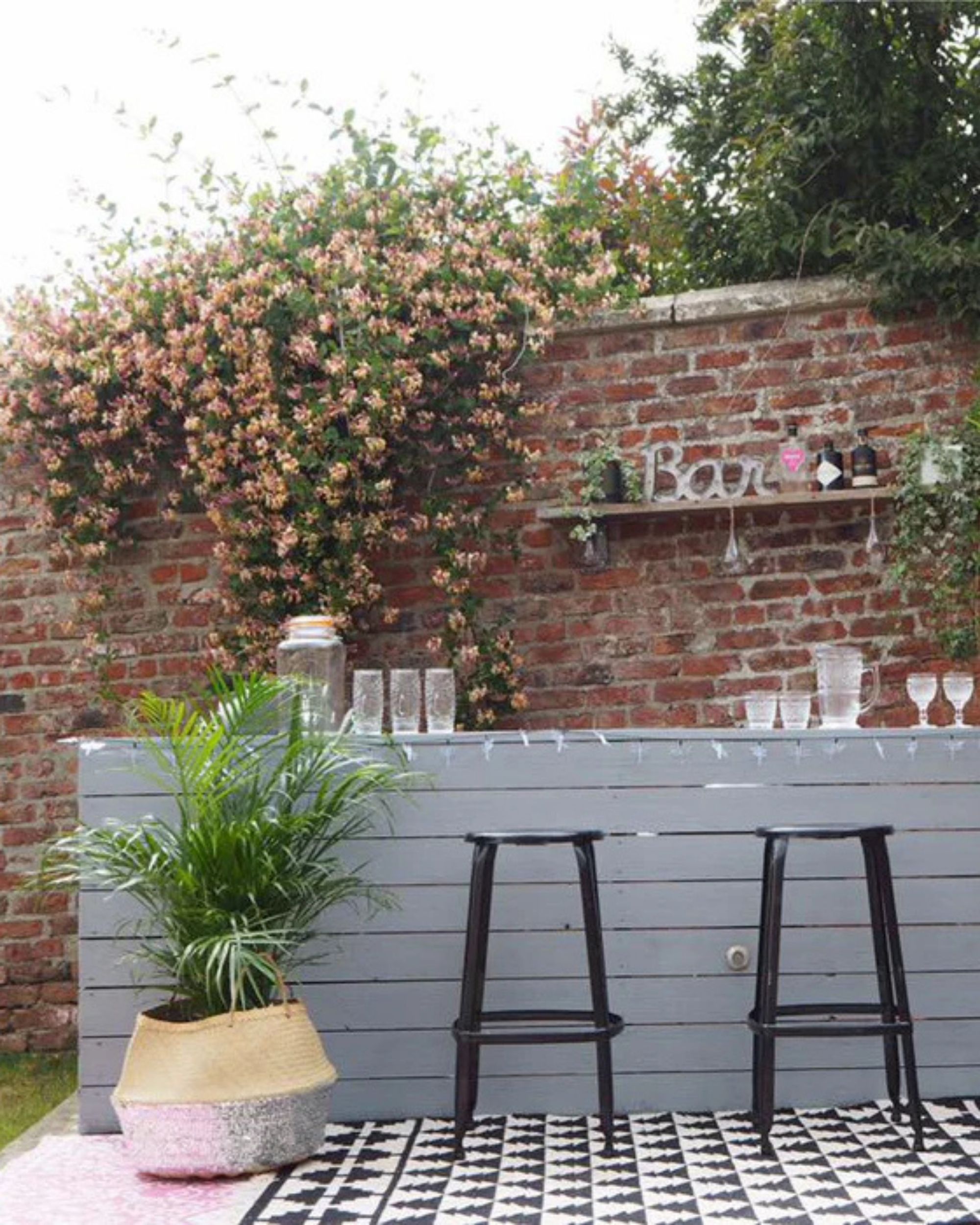 Image of a gray outdoor bar in front of a brick wall that has flowers growing over it. There are two black bar stools in front of the bar and a plant in a rattan basket.