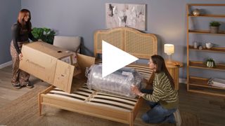 The image shows a screen shot of a video of two mattress testers in the process of unboxing a DreamCloud mattress in a neutrally decorated bedroom