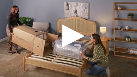 The image shows a screen shot of a video of two mattress testers in the process of unboxing a DreamCloud mattress in a neutrally decorated bedroom