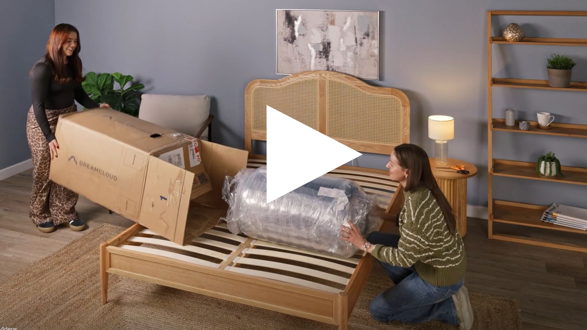 The image shows a screen shot of a video of two mattress testers in the process of unboxing a DreamCloud mattress in a neutrally decorated bedroom