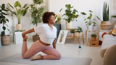 woman in a living room setting doing a hip flexor pilates exercise on an exercise mat with lots of indoor plants behind her. 