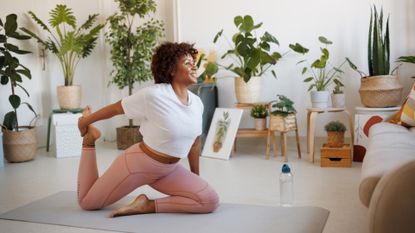 woman in a living room setting doing a hip flexor pilates exercise on an exercise mat with lots of indoor plants behind her.