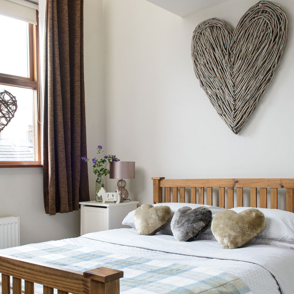 Small white bedroom with wooden bed brown curtains and wicker heart wall decor