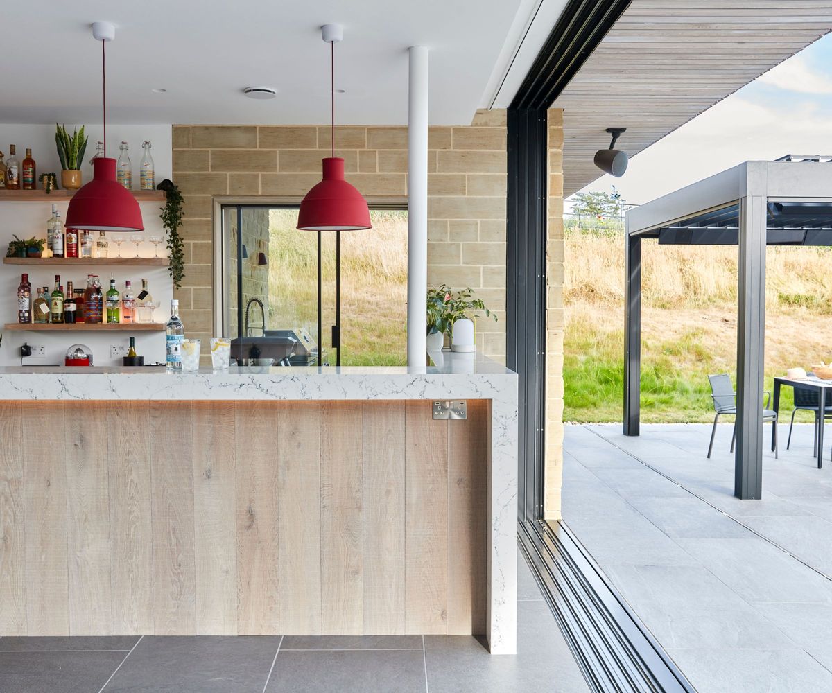 A pool house bar interior with a marble and oak island, two red pendant lights, open shelves of spirits, sliding glazed doors opening to a slate-tiled terrace with a pergola.
