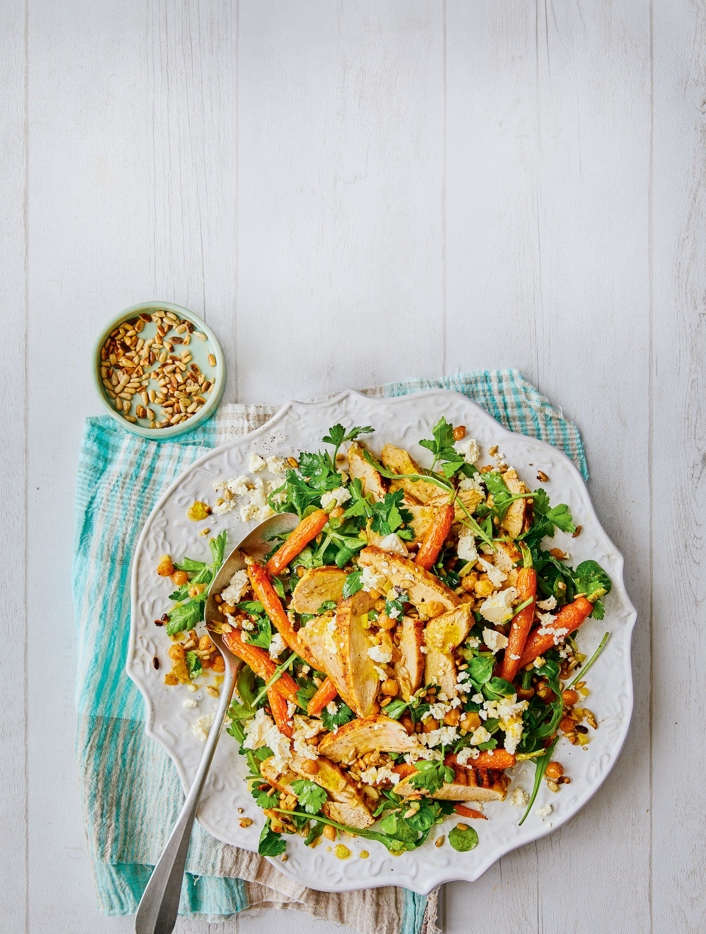A vibrant salad featuring mixed greens, sliced chicken, baby carrots, and crumbled cheese, served on a decorative white plate with a small bowl of pine nuts on the side.