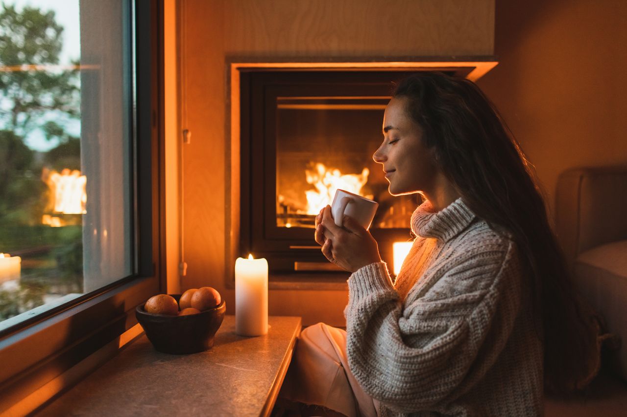 A happy woman sat in front of a warm fire and a lit candle, holding a mug of tea with her eyes closed.