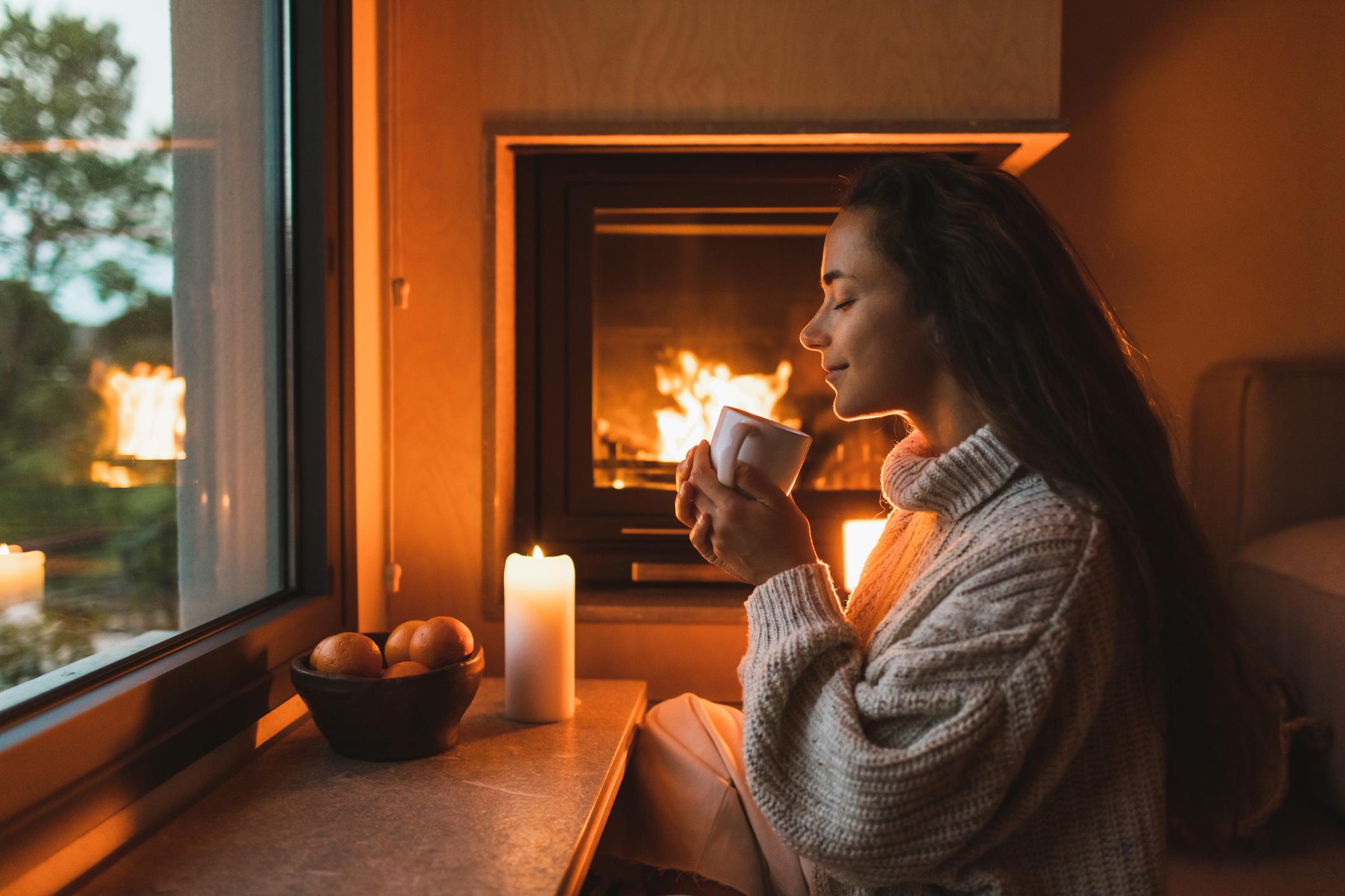  A happy woman sat in front of a warm fire and a lit candle, holding a mug of tea with her eyes closed. 