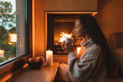 A happy woman sat in front of a warm fire and a lit candle, holding a mug of tea with her eyes closed.