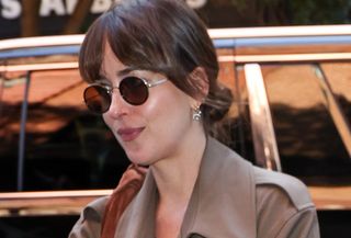 Dakota Johnson wearing a brown trench coat and jeans with a brown suede slouchy bag.