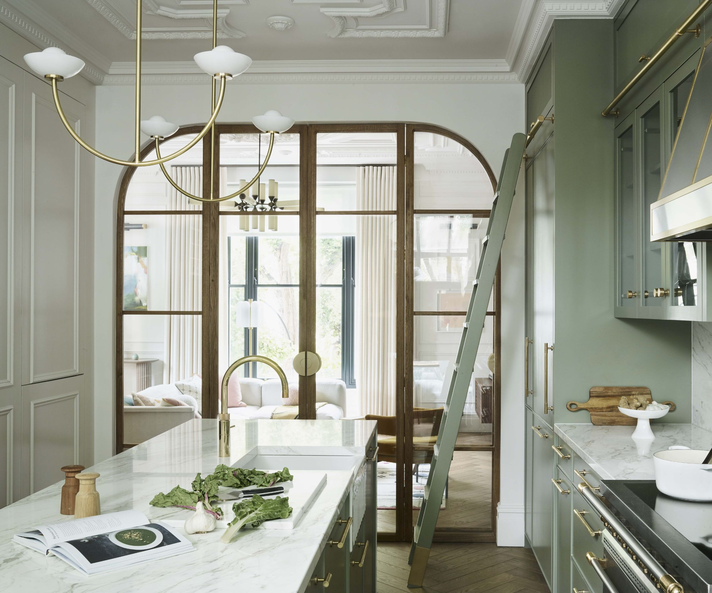 A kitchen with green cabinets, a wooden kitchen island, marble countertops, and glass doors separating the kitchen from the living space