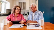 An older couple smile at each other as they work on financial planning at their kitchen table.