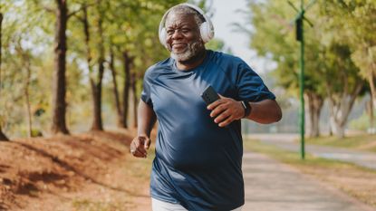 A smiling man in a t-shirt runs on a sidewalk, with trees lining either side. He wears over-ear headphones and a sports watch and carries his phone in his hand. 