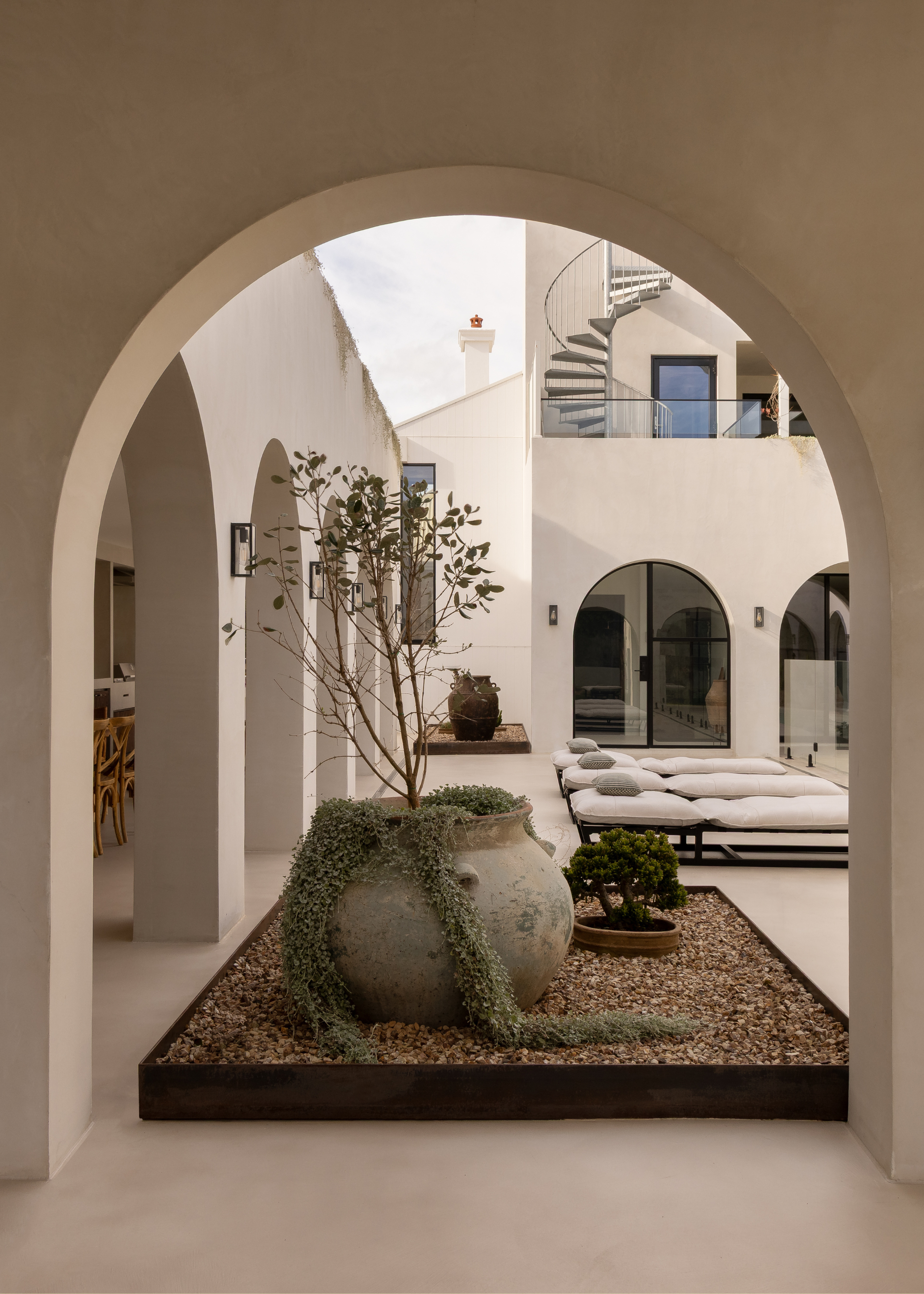 An outdoor lounging space with arched doorways leading to a small elevated stone bed with potted plants and day beds to the side