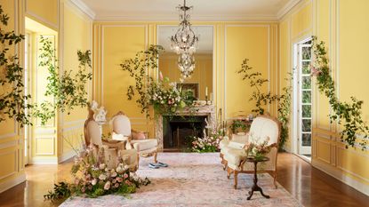 Bridgerton design ideas are so chic. Here is a yellow living room with a glass chandelier, three gold and cream chairs, a pink and blue floral rug, and flowers and vines on the floor and walls
