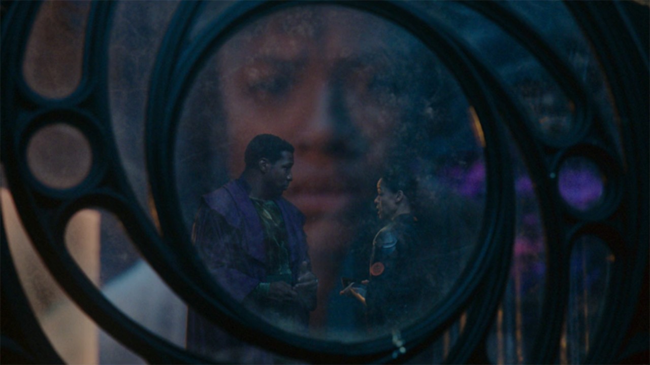 Image from the Marvel T.V. show Loki, season 2 episode 4. A man and woman standing in front of a large circular window are facing each other. There is a reflection of a woman's face in the glass window.