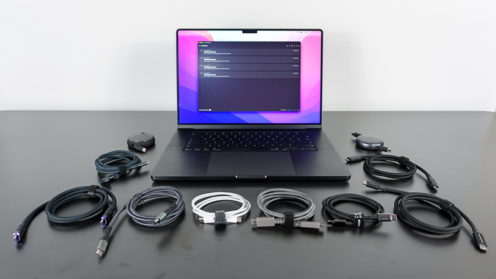 A MacBook Pro running the WhatCable app on a desk with 10 different USB-C to USB-C cables laid out in front of it