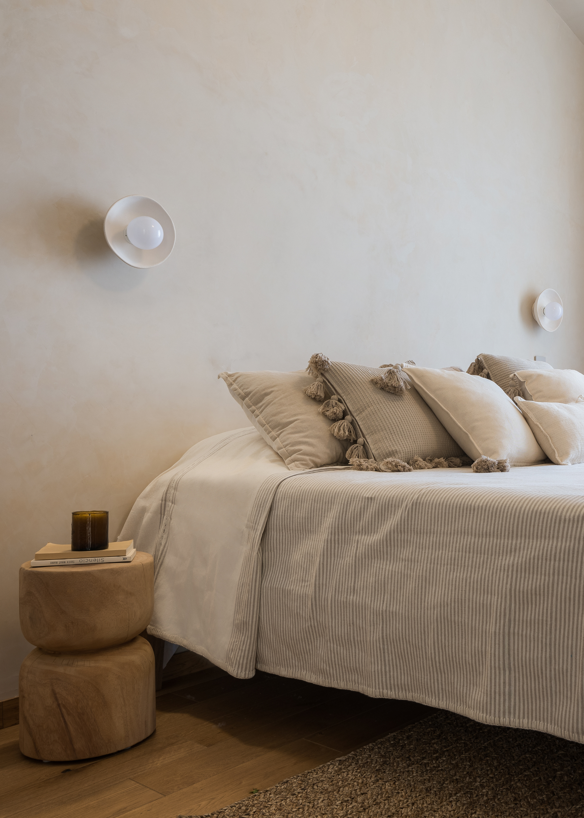 A beige bedroom with shelled wall sconces, a wood side table, a candle, a book, and a striped duvet with tasseled pillows