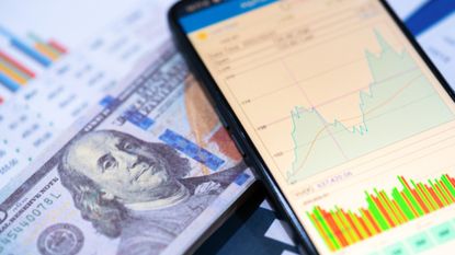 smartphone with a stock chart pulled up on the screen placed on top of a one-hundred dollar bill and financial charts