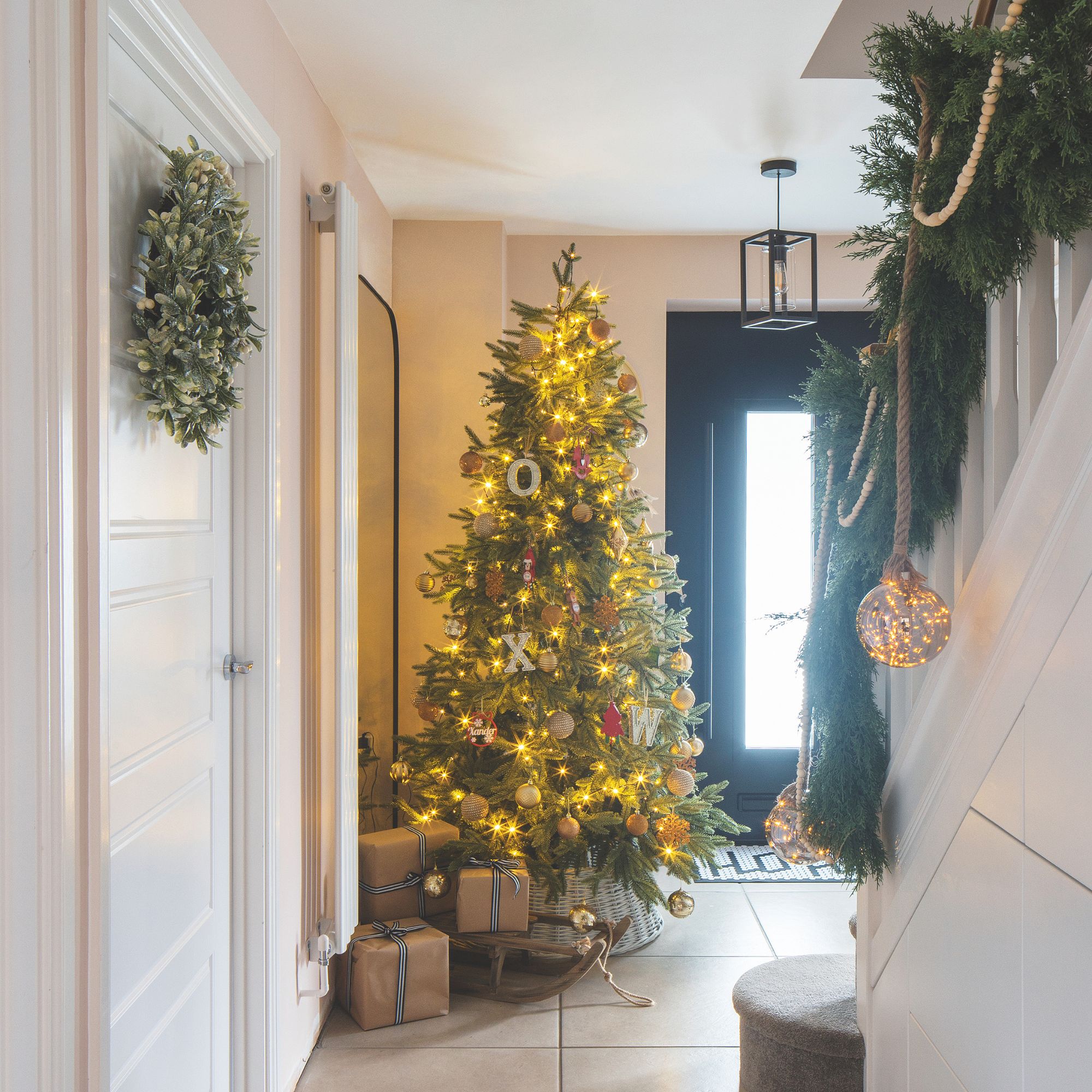 Decorated Christmas tree in a hallway with wrapped presents and sleigh decoration beneath it. There is also a garland going up the stairs and a wreath hanging on an internal door