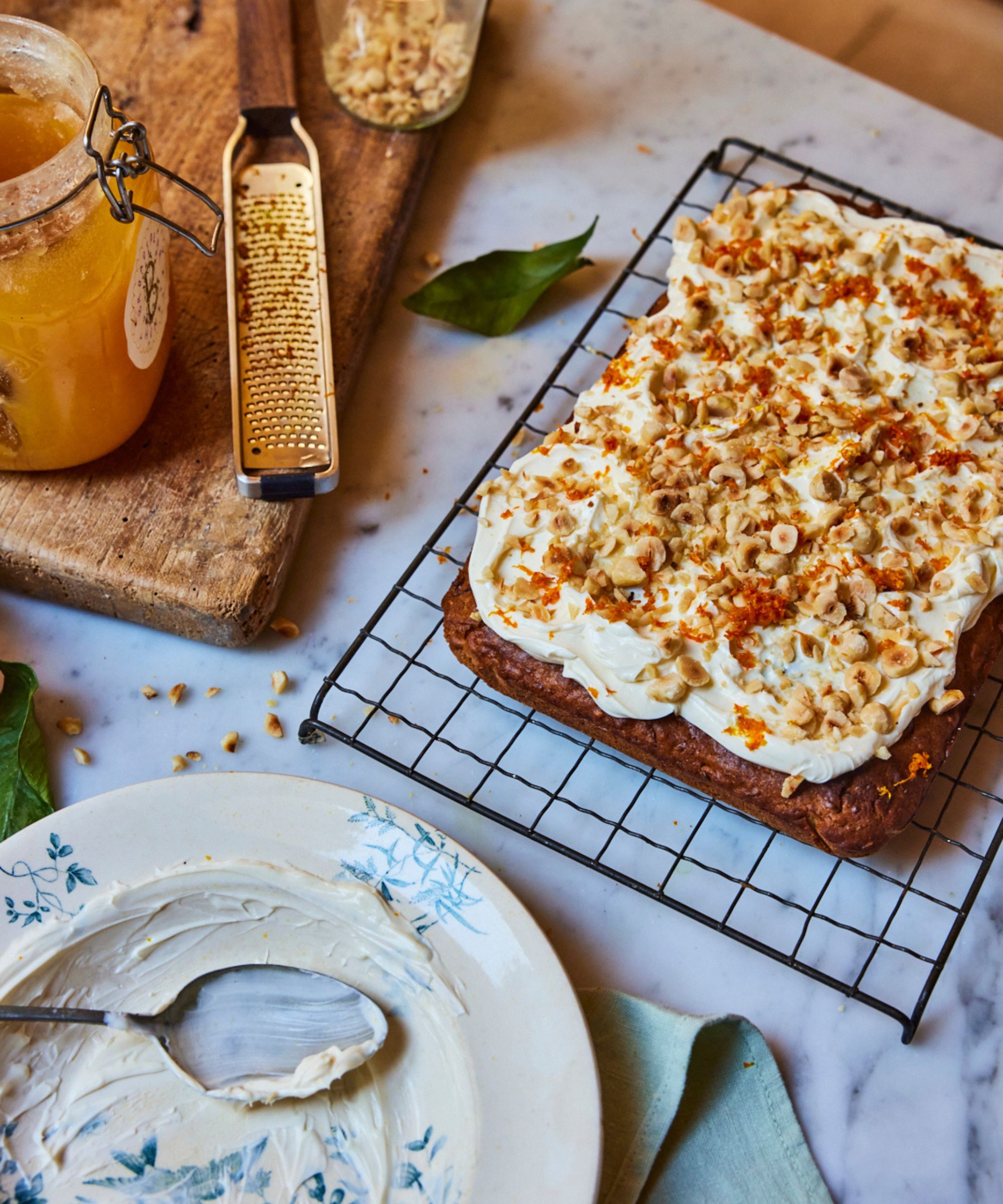 Parsnip cake with white frosting and nutty topping on a wire rack, next to a plate and spoon, wooden board with zester and pot of honey