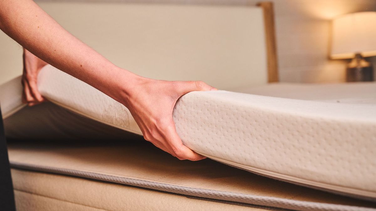 A woman places a thick latex mattress topper onto an older mattress to help it feel more comfortable and firm again