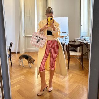 American influencer and fashion illustrator Jeny Walton wears a yellow trench coat, pink gingham Prada tote bag, red gingham Capri pants, and raffia Saint Laurent open-toe heeled mules