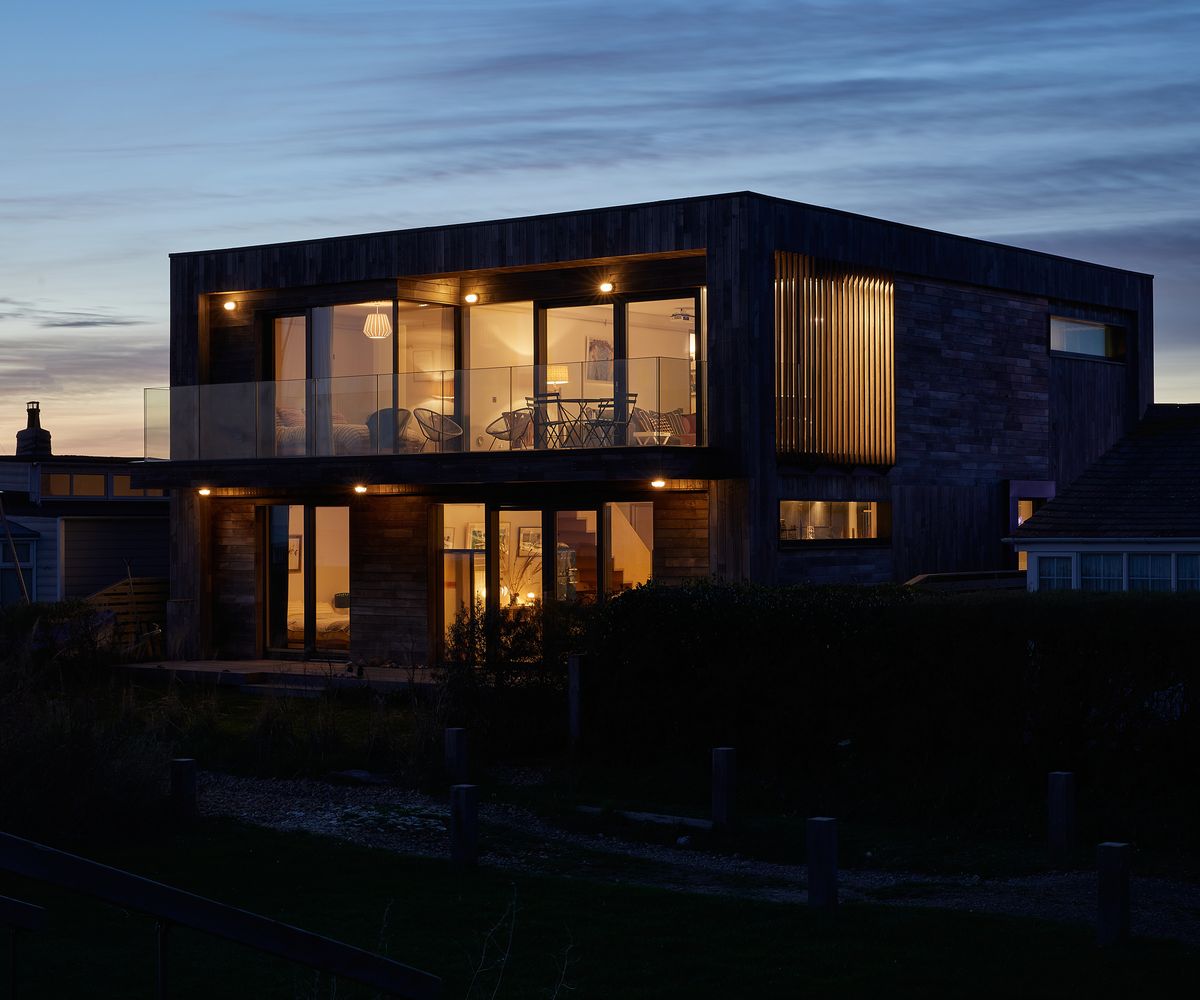 A contemporary seaside property at dusk with large windows and the lights on inside the house