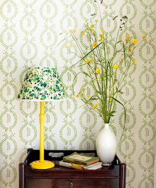 table against a green and white wallpaper with a yellow lamp and green and cream patterned shade, books and white vase with buttercups and grasses.