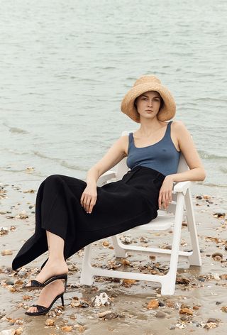 a photo of a woman on the beach sitting wearing a raffia hat with blue tank top and black trousers and black mules from the minimal shoe brand, dear frances