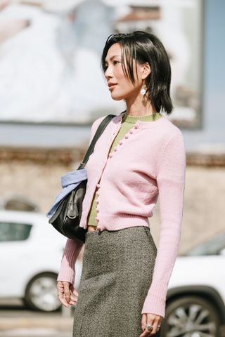 A woman wearing a fall outfit idea of a green sweater, pink cardigan, gray skirt, and black bag at Paris Fashion Week.