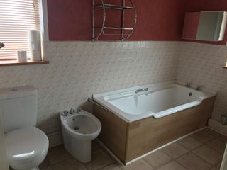 Bathroom makeover: before picture showing rectangular panelled bath, stone floor, old-fashioned floral tiles tiled halfway up the wall, and patterned red wallpaper above the tiles