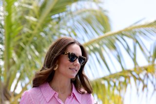 A shoulders up photo of Kate Middleton wearing a pink dress and sunglasses with a palm tree behind her