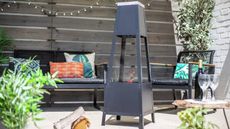 one of the best chiminea options in a garden with a wood and white brick wall, with black garden furniture and colourful cushions, and plants and fairy lights 