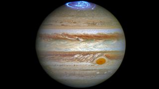 Jupiter against the black backdrop of space. Horizontal bands of orange, brown and beige cover the planet. Blue ribbons of light are auroras at the top of the planet and the Great Red Spot can be seen towards the lower right of the image as a rusty red circle.