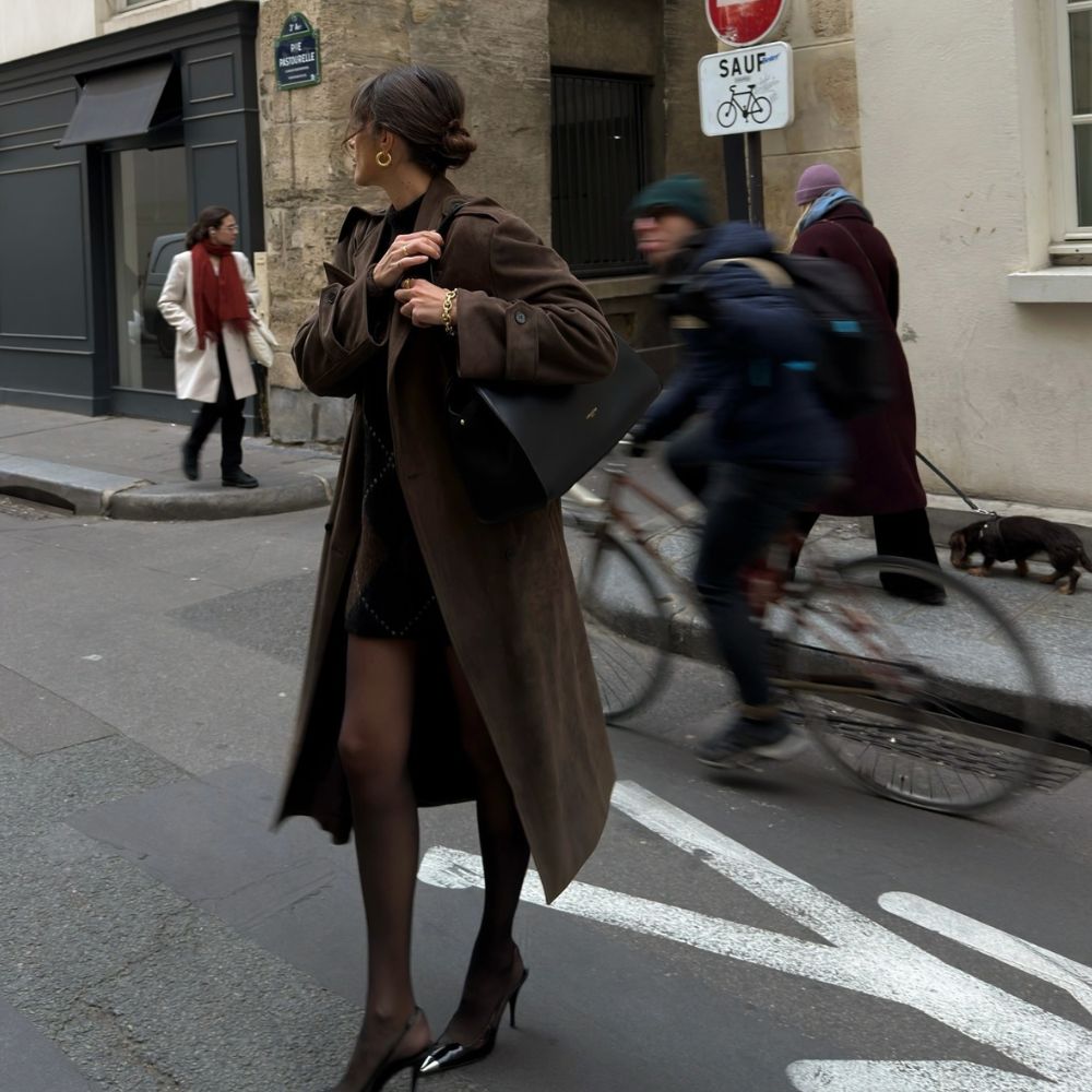 French influencer @juliesfi walks down the streets of Paris wearing a dark brown trench coat with tights and slingback heels.
