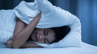 A woman holding a pillow over head head at night as she struggles to sleep from noise