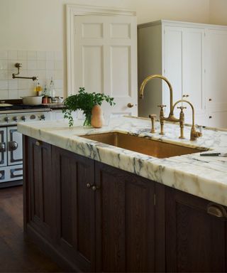 A traditional kitchen with a dark wood island, veined marble worktop, and an aged brass sink. A pot filler tap and a classic range cooker enhance the vintage aesthetic.