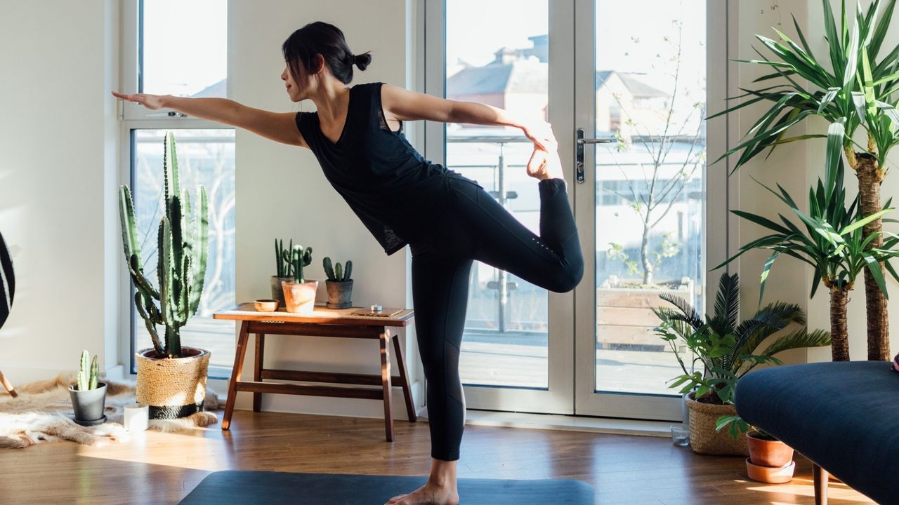 woman wearing black vest and leggings doing a one legged balance yoga pose in living room setting with windows and plants behind her