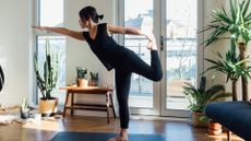 woman wearing black vest and leggings doing a one legged balance yoga pose in living room setting with windows and plants behind her